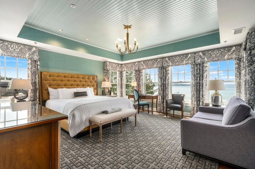 Bright hotel room with a king bed, large windows, and a view of the water, featuring elegant decor and cozy seating.