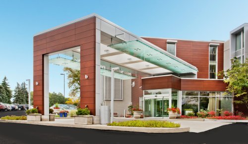 Modern building entrance with glass canopy, wooden accents, and landscaped planters under a clear blue sky.
