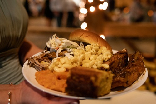 A plate of food featuring pulled pork, coleslaw, macaroni and cheese, cornbread, and a bun, held by a person.