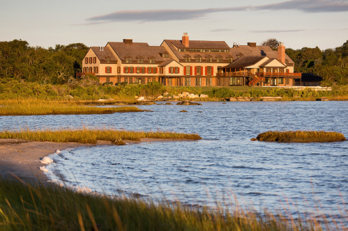 A large, elegant building with red shutters sits by a calm waterway, surrounded by greenery and sandy shores.