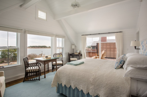 Bright, airy bedroom with a large bed, desk, and views of a lake through large windows. Cozy coastal decor.