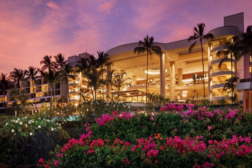 A luxurious hotel illuminated at dusk, surrounded by vibrant pink flowers and palm trees against a colorful sky.