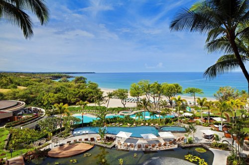 A scenic view of a resort with pools, palm trees, and a beach under a clear blue sky.