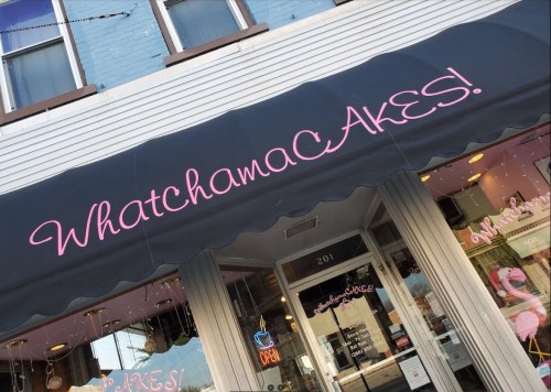 Sign for "WhatchamaCakes!" bakery with a pink awning and storefront, featuring an open sign.