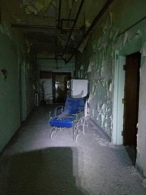 Dimly lit hallway with peeling paint, featuring an abandoned blue wheelchair and a refrigerator.