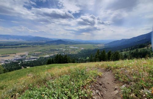 A scenic view from a hillside overlooking a valley, with mountains in the distance and a partly cloudy sky.