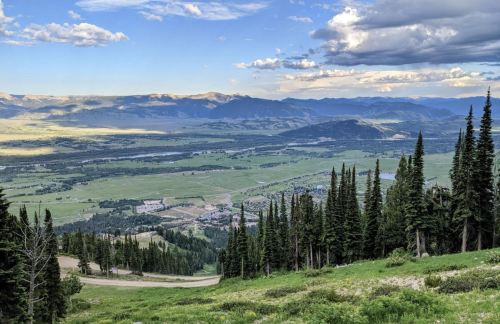 A panoramic view of a lush green valley surrounded by mountains, with scattered clouds in a blue sky.