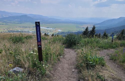 A scenic view from a hiking trail, with a signpost and lush greenery overlooking a valley and distant mountains.
