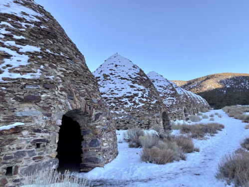 Stone kilns covered in snow, with a clear blue sky and mountains in the background. Pathway leads through the scene.