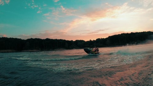 A person rides a jet ski on a lake at sunset, with vibrant colors in the sky and trees lining the shore.