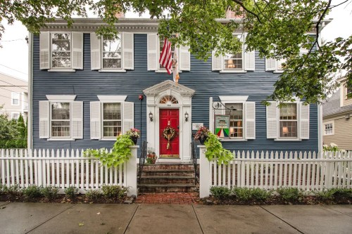 A blue house with white shutters, a red door, and a picket fence, adorned with plants and an American flag.