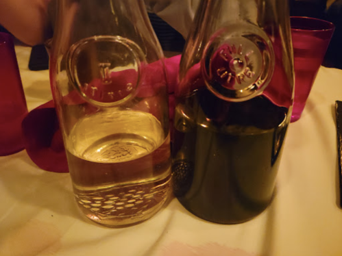 Two glass bottles on a table, one filled with clear liquid and the other with dark liquid, beside red cups.