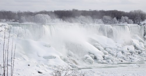 A snowy landscape featuring a frozen waterfall, with mist rising and trees in the background.