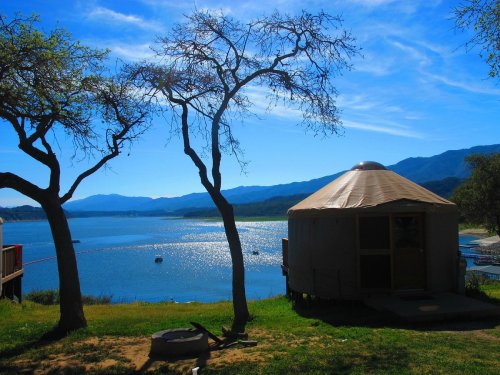 A serene lakeside scene featuring a yurt, trees, and mountains under a clear blue sky.