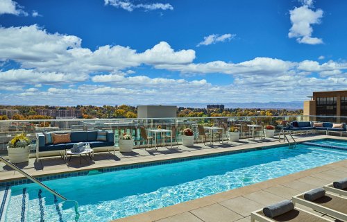 Rooftop pool with lounge chairs and tables, surrounded by a scenic view of trees and clouds under a blue sky.