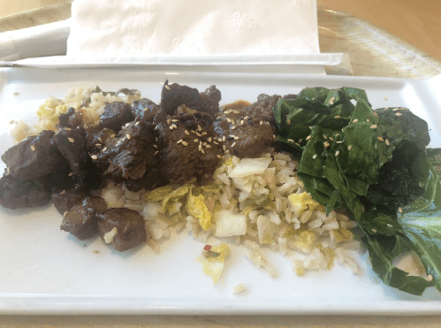 A plate of beef stir-fry with rice, sautéed greens, and sesame seeds, served on a white dish.