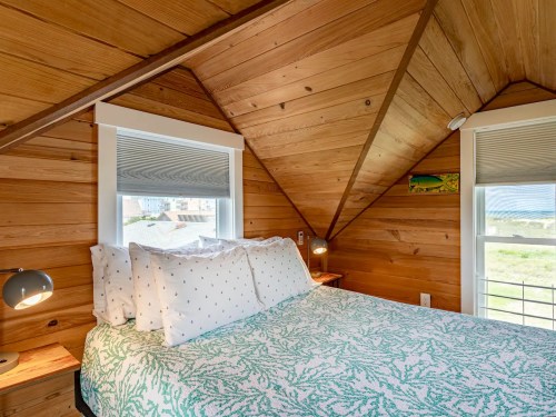 Cozy bedroom with wooden walls, a green patterned bedspread, and large windows letting in natural light.