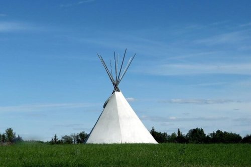 A white teepee stands on green grass under a clear blue sky with a few clouds.
