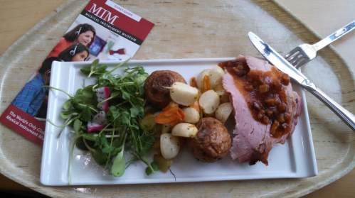 A plate of sliced ham with sauce, roasted vegetables, and a salad, alongside a brochure for the Musical Instrument Museum.