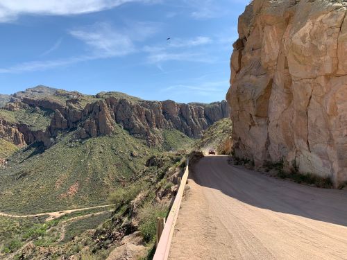 A winding dirt road alongside a rocky cliff, with lush green mountains in the background under a clear blue sky.