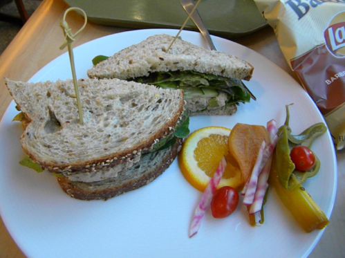 A sandwich on whole grain bread with greens, pickles, and a side of colorful vegetables and chips on a plate.