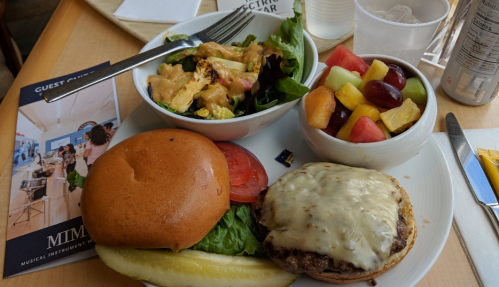 A plate with a cheeseburger, lettuce, tomato, pickle, salad, and a bowl of mixed fruit.