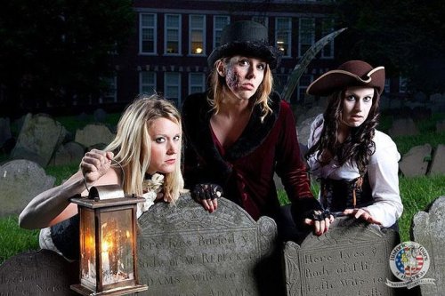 Three women in vintage costumes pose dramatically in a graveyard, holding a lantern and surrounded by tombstones.