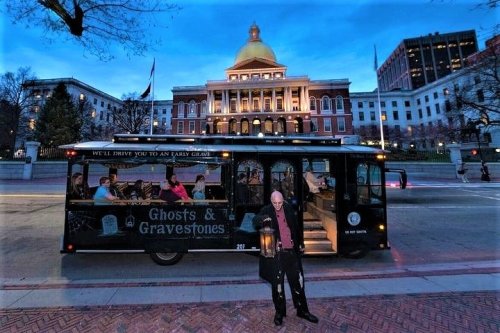 A man holding a lantern stands in front of a trolley labeled "Ghosts & Gravestones" near a historic building at dusk.