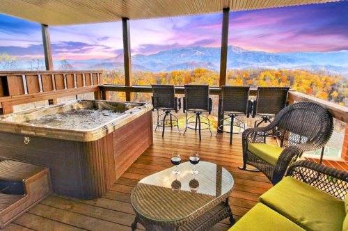 A cozy balcony with a hot tub, chairs, and a stunning view of mountains and autumn foliage at sunset.
