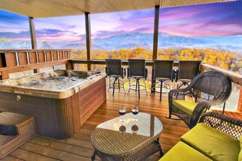 A cozy balcony with a hot tub, seating, and a stunning view of mountains and autumn foliage at sunset.