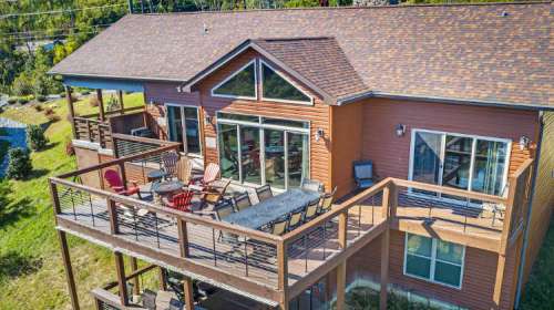 A modern house with a large deck, featuring outdoor seating and a fire pit, surrounded by greenery.