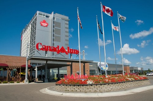 Exterior view of a Canada Inn hotel with flags and flower beds under a clear blue sky.