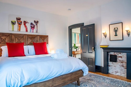 Cozy bedroom with a large bed, red pillows, artwork on the wall, and a decorative fireplace. Bright and inviting atmosphere.