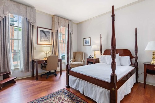 Cozy bedroom featuring a four-poster bed, desk, armchair, and warm wooden accents. Natural light from a window.