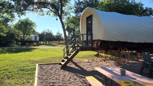 Two covered wagons on a grassy area, with a wooden picnic table in the foreground and trees in the background.