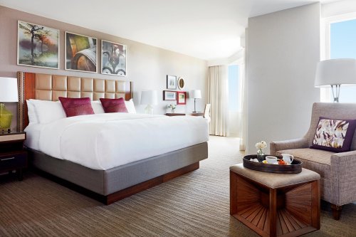 Modern hotel room featuring a king-sized bed, armchair, and a small table with refreshments, bright natural light.