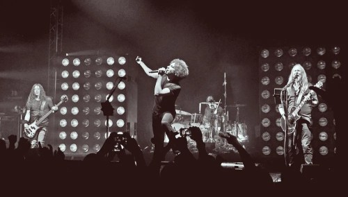 A rock band performs on stage, with a singer in front and musicians playing instruments, under dramatic lighting.