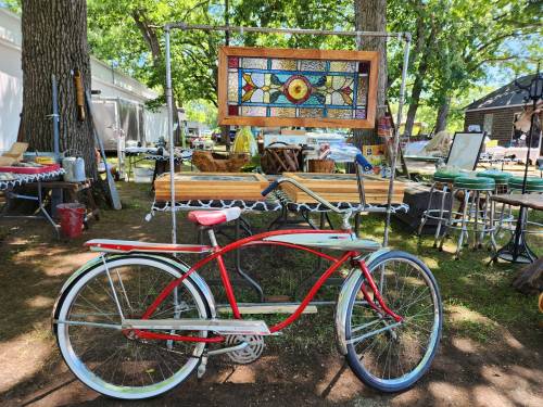 A vintage red bicycle stands beside a colorful stained glass piece in a sunny outdoor market setting.