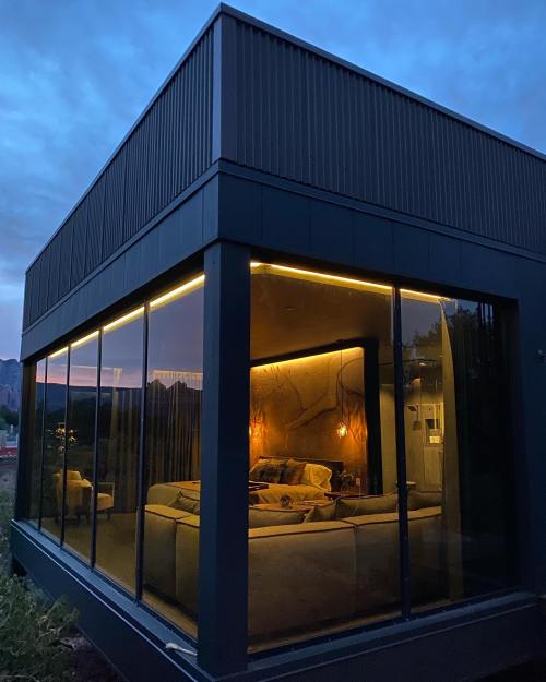 Modern glass-walled house at dusk, featuring warm interior lighting and scenic mountain views.