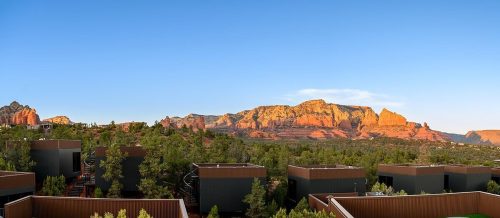 Panoramic view of red rock formations and green trees under a clear blue sky, with modern buildings in the foreground.