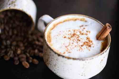 A cup of frothy coffee topped with cinnamon, surrounded by scattered coffee beans.