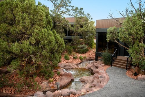 A serene landscape featuring a small stream, rocky terrain, and lush greenery near modern buildings.