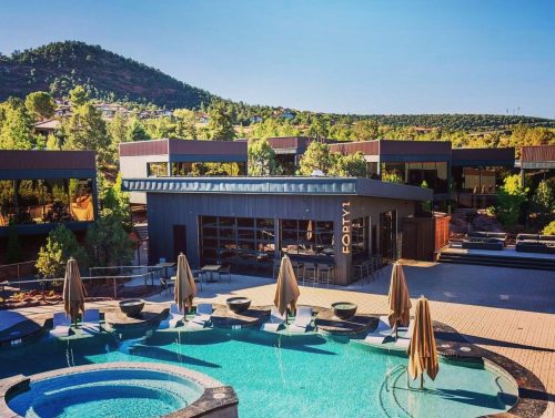 A modern hotel with a pool and lounge area, surrounded by trees and mountains under a clear blue sky.