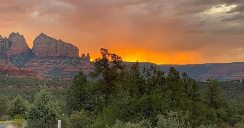 Sunset over red rock formations and green trees in a scenic landscape, with dramatic clouds in the sky.