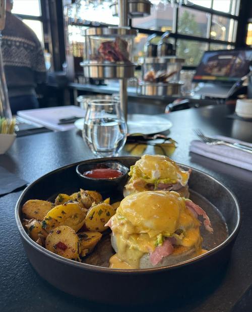 A plate of eggs Benedict with ham, served with seasoned potatoes and a side of sauce, on a restaurant table.