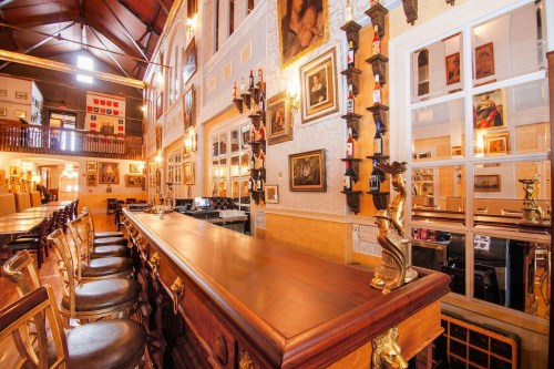 A warm, inviting bar interior with wooden counters, art on the walls, and high ceilings, creating a cozy atmosphere.