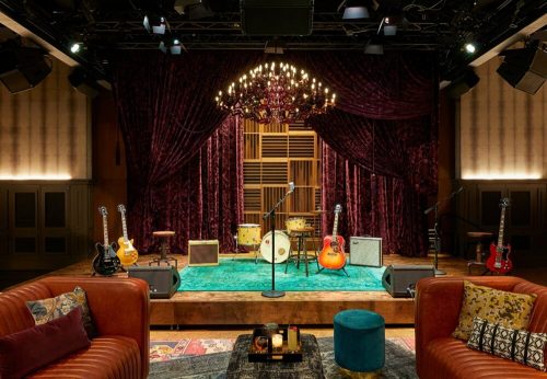 A cozy music stage with guitars, drums, and a chandelier, set against rich purple curtains and a teal rug.