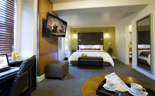 A modern hotel room featuring a bed, TV, desk, and seating area with a mirror and natural light from windows.