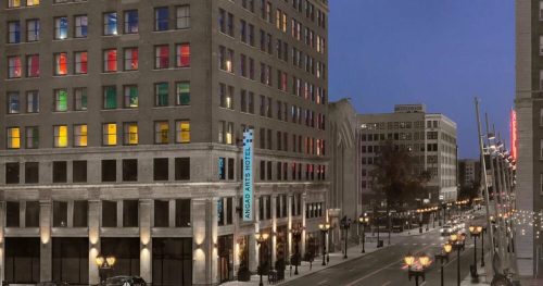 A city street at dusk featuring a hotel with colorful windows and nearby buildings illuminated by streetlights.