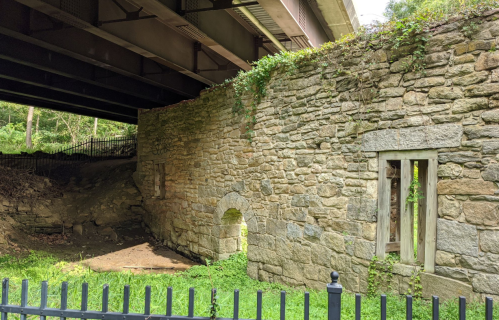 Stone wall ruins covered in greenery, located beneath a bridge, with a dirt area and a black fence nearby.